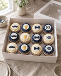 Stag Do Cupcakes: twelve cupcakes in white cases with navy and white buttercream swirls, rows of four, topped in turn with flat sugar discs: a black moustache, a pint of beer, a navy bow tie, and a white plaque reading MR in navy piped letters