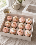 Milestone Number Cupcakes: twelve cupcakes in white cases with blush pink buttercream swirls; six are topped with a flat white sugar disc showing a gold number 30 edged in gold, the rest with gold sugar pearls