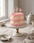 Ballerina Lambeth Cake: smooth peach-pink buttercream with cream drapes and pink ruffles, sugar pearls on the swags, and three small paper ballerina toppers in pink tutus standing on the top