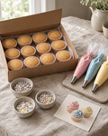 Decorate Your Own Cupcakes Kit: a kit laid out on the linen: twelve plain baked cupcakes in white cases in an open box, three filled piping bags of pink, blue and yellow buttercream with star nozzles, three small pots of sprinkles and sugar stars, and a printed recipe card