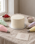 Pipe Your Own Lambeth Kit: a kit laid out on the linen: a smooth white crumb-coated 6 inch cake on a board, four filled piping bags of pastel buttercream (pink, mint, lilac, yellow) with star nozzles, a small pot of glacé cherries, a pot of sprinkles and a printed card reading PIPE YOUR OWN