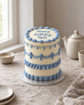 Cornflower Sampler Cake: a tall round ivory cake with four stacked bands of cornflower blue piping, each band a different style: fine string lace, swags with little piped bows, a ruffle, and a row of shells, with cornflower sugar pearls at the swag points