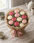 Cupcake Bouquet: a bouquet of twelve cupcakes piped as roses in blush pink, deep pink and cream, arranged tightly in a round bouquet shape with piped green leaves between them, wrapped in kraft paper and tied with a pink satin ribbon, standing on the linen