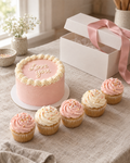 Bento and Cupcakes Gift Box: an open white gift box holding a 4 inch bento cake in blush buttercream with a cream ruffled border, next to five cupcakes with blush and cream swirls in a row, a pink satin ribbon draped over the open lid